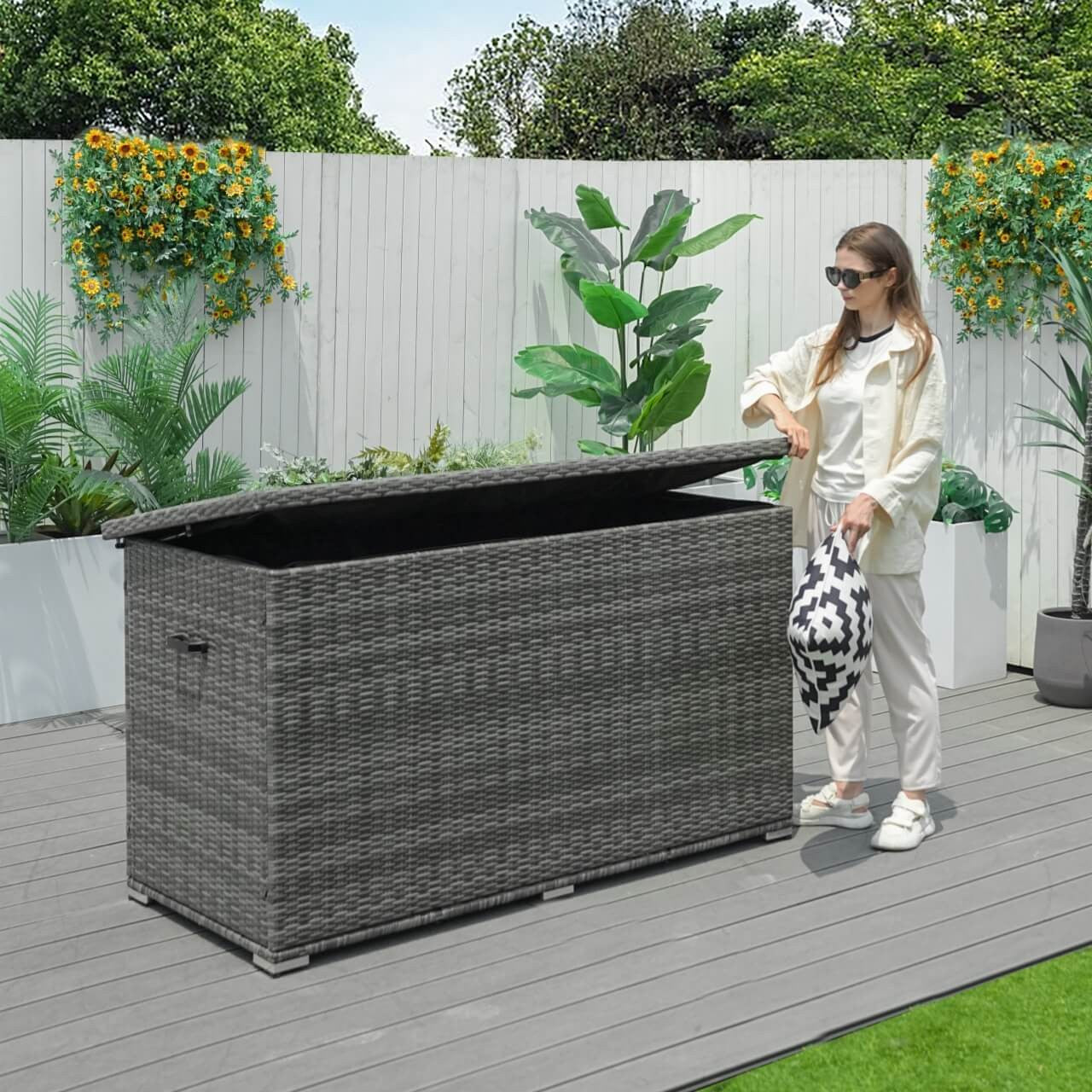 A woman is opening the lid of a gray outdoor wicker storage box placed on a wooden deck with a garden in the background.
