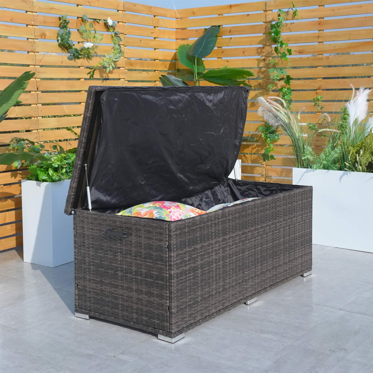 A medium iron rattan storage box with a black waterproof inner liner, placed on a patio next to a wooden fence and plants. The box appears to be closed, with a handle visible on the side.