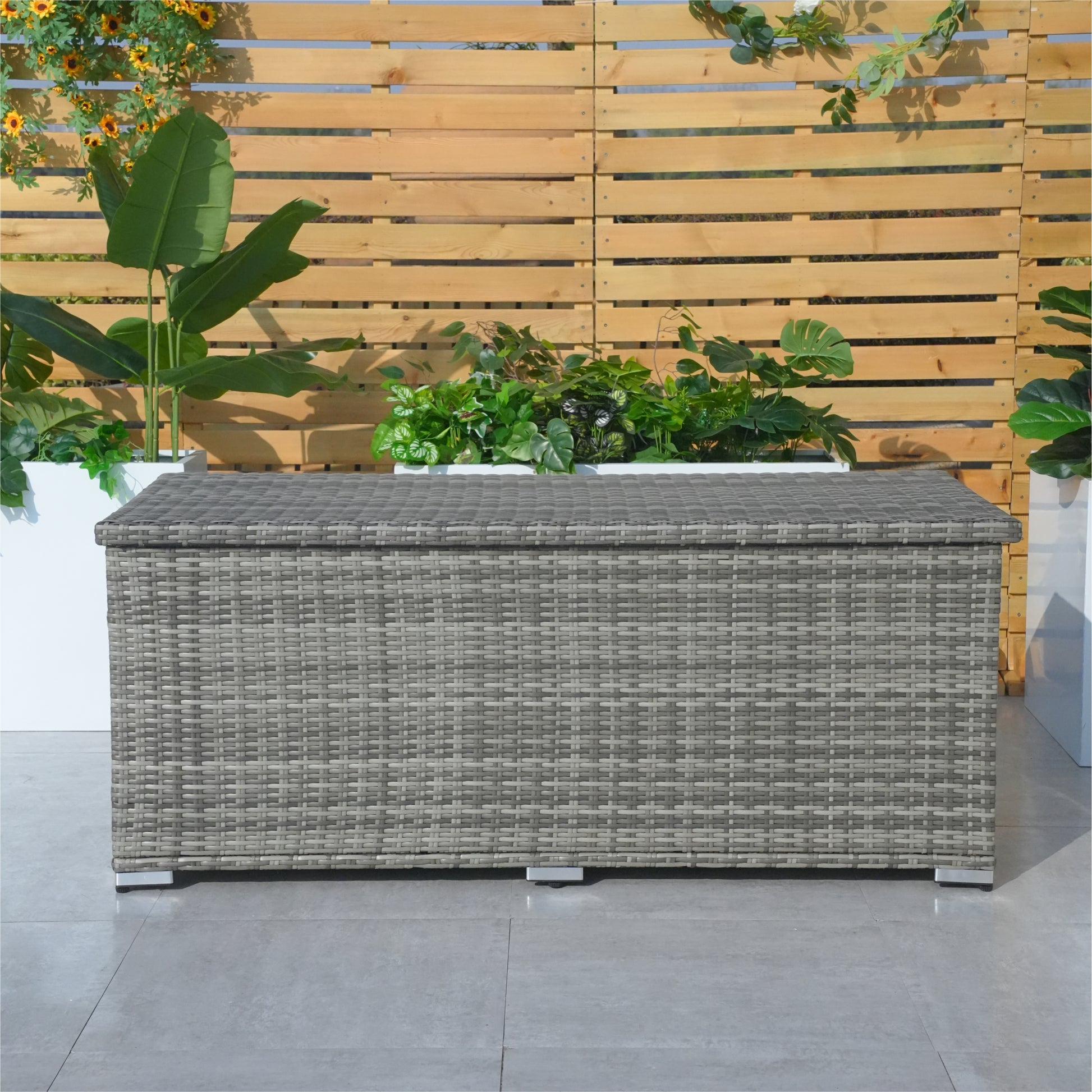 A medium iron rattan storage box with a black waterproof inner liner, placed on a patio next to a wooden fence and plants. The box appears to be closed, with a handle visible on the side.