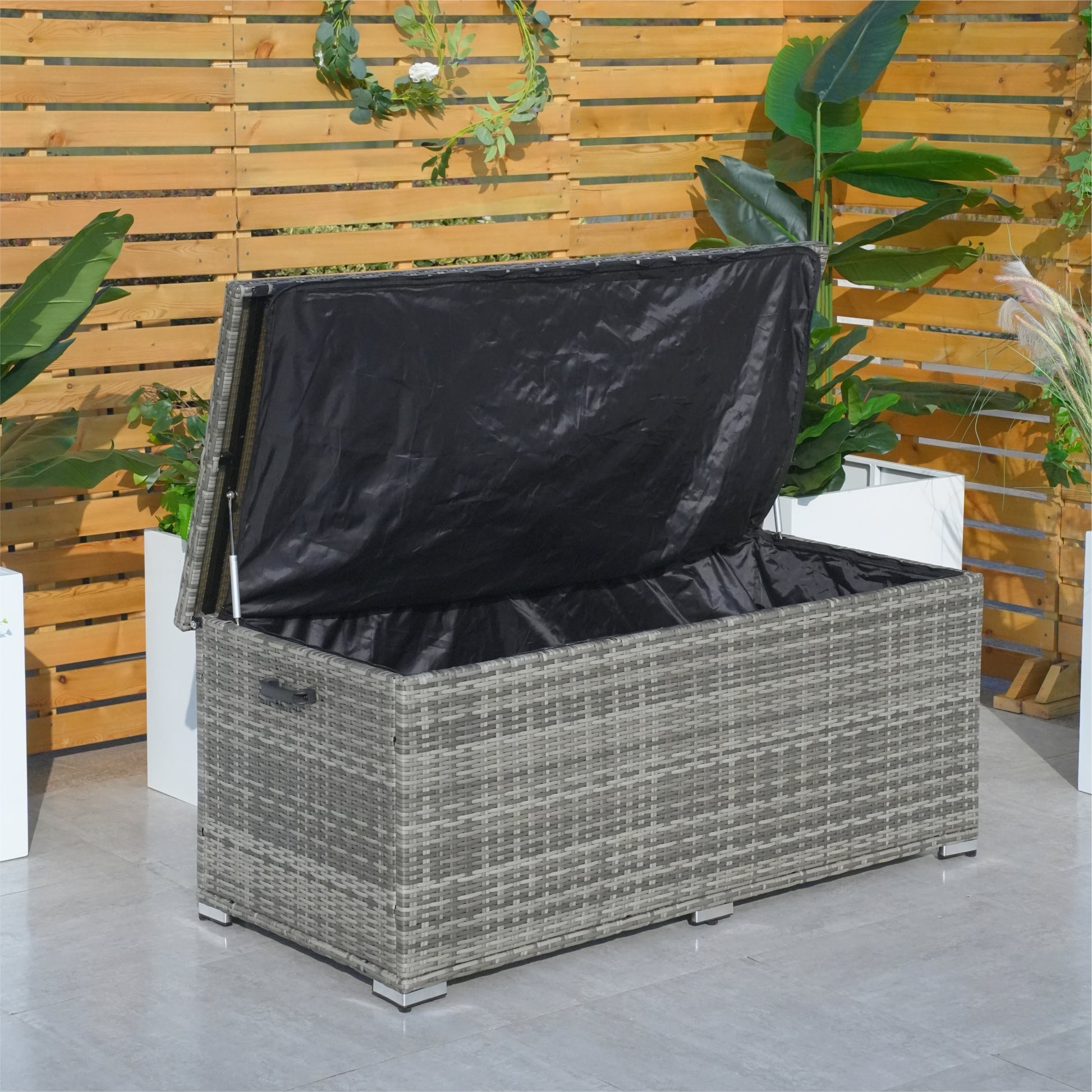 A compact aluminium rattan storage box with a black waterproof cover, placed on a patio between two potted plants.