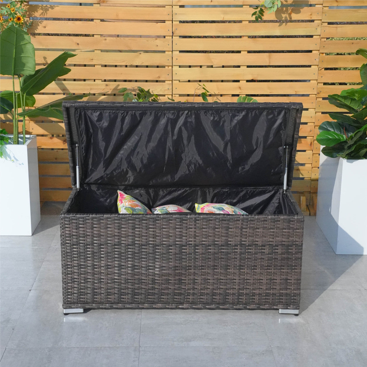 A compact aluminium rattan storage box with a black waterproof cover, placed on a patio between two potted plants.