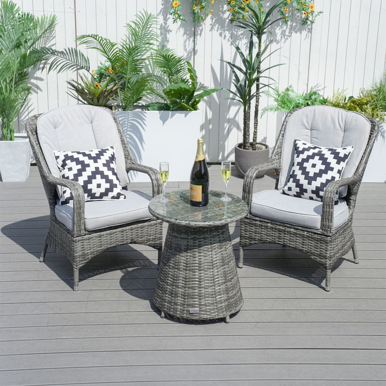 A 3-piece outdoor bistro set with two cushioned chairs and a round wicker table with a glass top, set on a gray outdoor patio with plants in the background.
