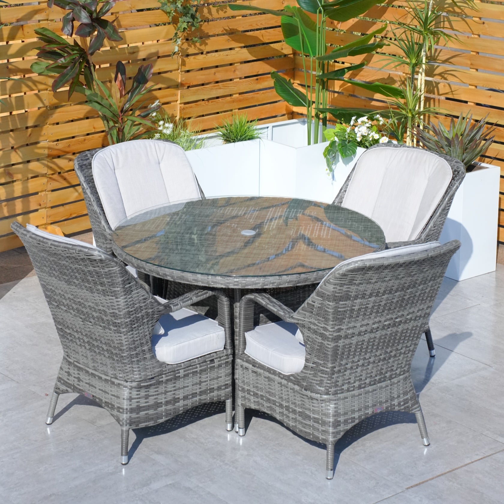 A Parma 4-Seat Outdoor Dining Set with a round table and four chairs with cushions, placed on a deck surrounded by green plants.