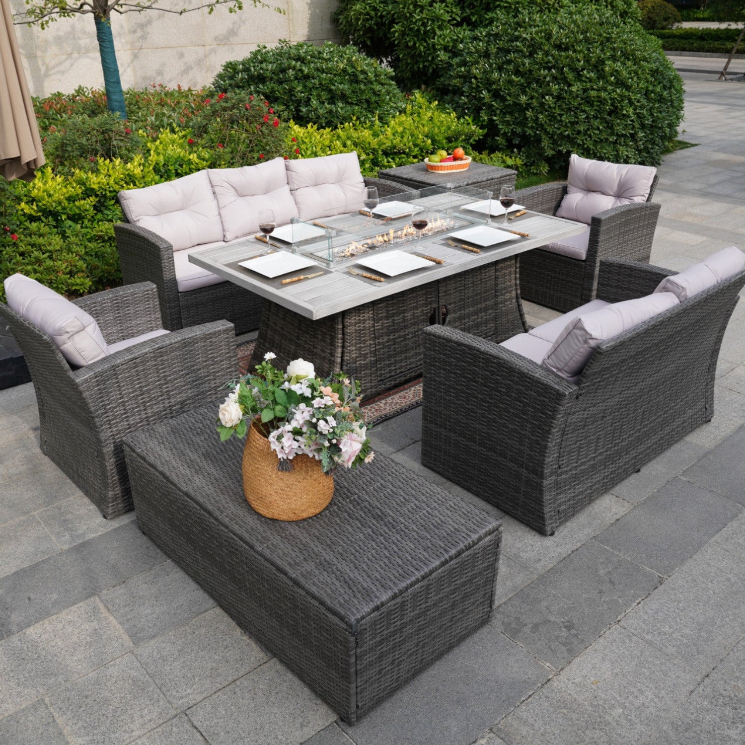 Outdoor patio furniture set including a fire pit table, a three-seat sofa, a double sofa, and single sofas around it, all with grey cushions, alongside a coffee table with built-in storage and a side table with built-in storage. The set is placed on a paved outdoor area with greenery in the background.