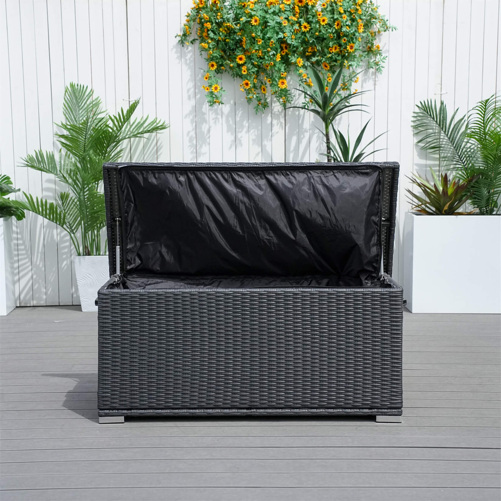 Wicker storage bench with black cover on a wooden deck with plants in the background