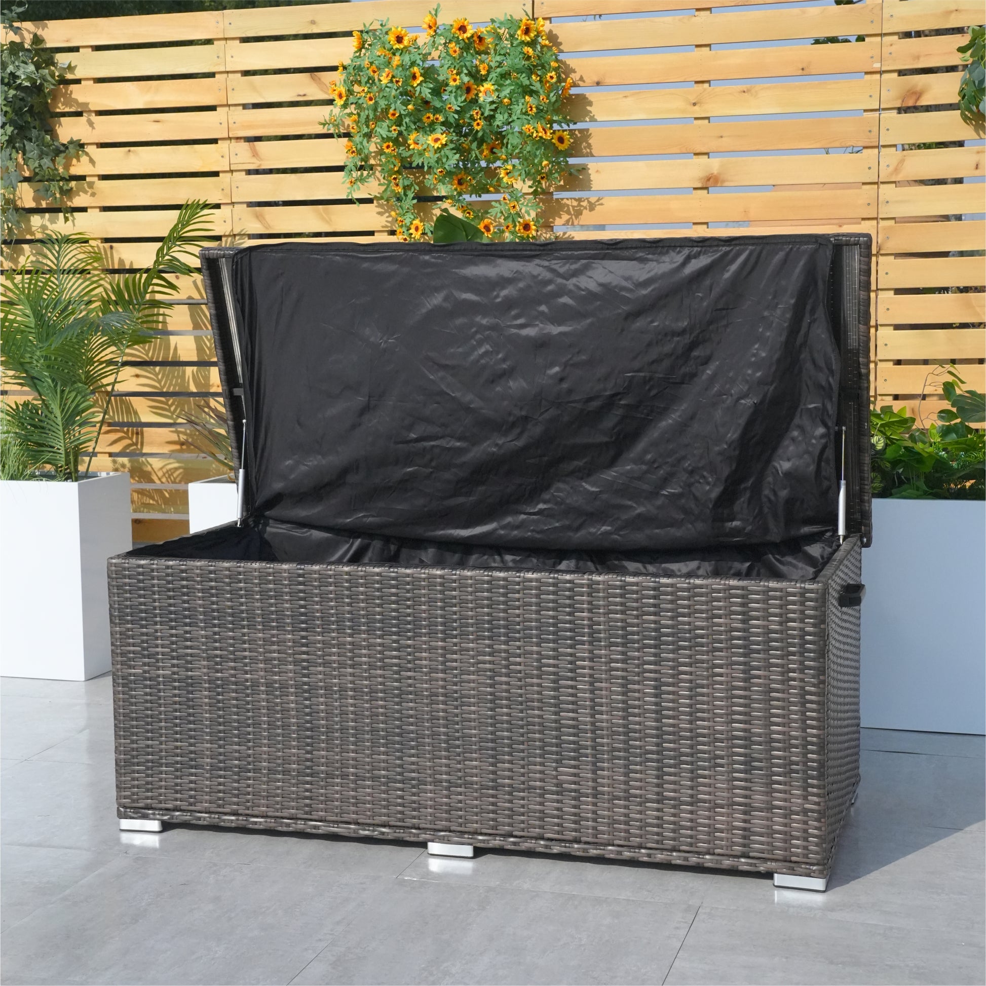 A compact aluminium rattan storage box with a black waterproof cover, placed on a patio between two potted plants.